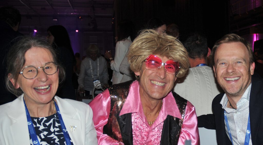 Typetje fotograaf poseert voor zijn eigen camera met twee lachende gasten op een internationaal evenement.