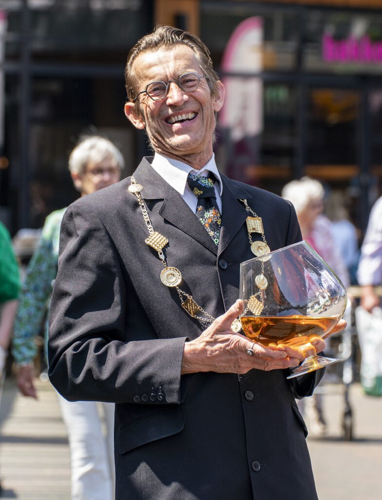 Typetje burgemeester van Free Fantasy lachend met groot glas cognac op evenement.