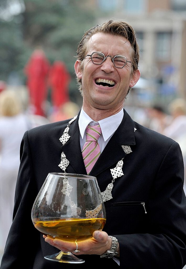 Typetje burgemeester lachend met groot glas cognac op straattheaterdag.